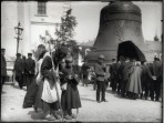 A bearded man with a sack and a long stick leans over next to a young boy while a queue of people form next to a large bell behind