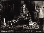 A seated woman with a paterned scarf around her head and shoulders, her legs stretched out in front of her and her arms laid open on the floor sits with her mouth open and her head turned up towards the ceiling