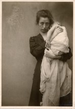 Woman with dark hair and a melancholic expression holds a baby wrapped in a white cloth up to her shoulder