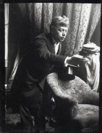 Model in a dark suit holding a top hat and leaning forward over an armchair with his right hand held out in front of him
