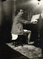 Gauguin sitting at a harmonium wearing only a shirt and jacket, turning towards the camera