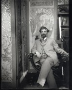 Mucha dressed in a suit seated in an ornate chair in front of 3 Sarah Bernhardt posters, looking at the camera