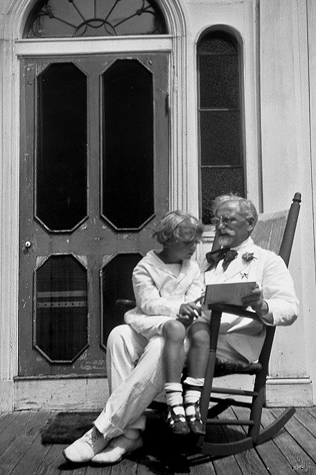 Mucha dressed in a white suit sitting on a rocking chair on a wooden veranda with his son Jiří on his knee