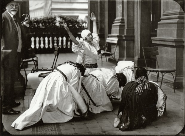 Mucha looks on as 4 women in traditional costume crouch down on the floor in a huddle and a 5th woman with a white headdress holds her arms up to the sky