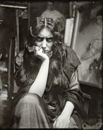 A seated female model with bedraggled long, dark hair and an angry expression leans on her knee with her head on her hand