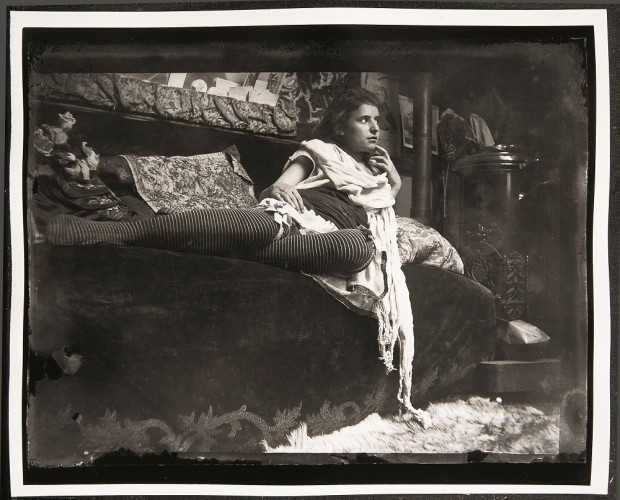 Young woman in stripey stockings with a white scarf around her neck reclining on a velvet couch next to a stove.