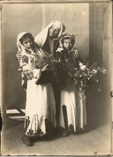A woman in a white headdress stands with her arms around two girls in embroidered tunics holding bunches of flowers