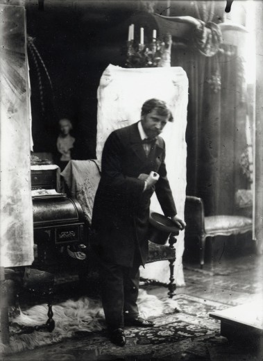 Mucha dressed in formal clothes holding a top hat and leaning forward with his left hand on a chair behind him