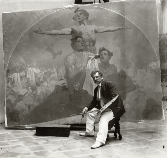 Mucha seated on a stool in front of his mural with a paint palette and paint brush in his hand