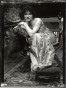 Bare-shouldered model sitting on an ornate wooden chair and leaning on a cushion placed on the right arm of the chair