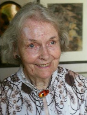 Geraldine Mucha Front facing portrait of Geraldine Mucha with a patterned blouse and a broach sitting in front of two of Mucha's works