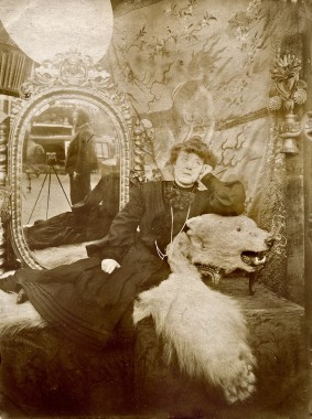 Mucha taking a picture of a model, rue Val de Grâce Model reclining against the head of a bear skin rug with an oval mirror behind revealing the reflection of Mucha with his camera