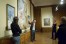 A man points at Mucha's 'Madonna of the Lilies' aa he turns towards a group of visitors in a wood-paneled exhibition space